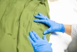 Hands with blue protective gloves are spreading apart some green fabric to show bullet holes in the fabric.