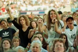 A crowd of women, viewing some sort of entertainment off-camera. Many are smiling and some have their arms raised. A larger crowd can be seen seated in a stadium behind them.