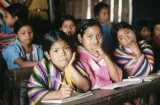 Children sit in a classroom with booklets in front of them on their desk. Three girls sitting close to the camera are looking directly into the camera. 