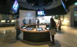 Six people in a museum gallery stand around a circular counter with video screens both embedded in the counter and hanging above them.