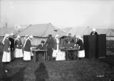 A black-and-white photograph of nurses and men in soldiers’ uniforms in front of a set of tents. To the right of the image, a nurse stands behind a voting screen. Only her head and shoulders can be seen.