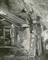 Two miners wearing helmets work underground in a uranium mine. One stands on a ladder, as the other miner watches him.