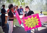 Two women are holding posters that advertise a “Plate sale fundraiser.” The woman on the viewer’s right is speaking into a flip phone that she is holding in front of her face, rather than up to her ear.