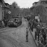 A crowd gathered on sidewalks, watching armoured vehicles driving through the cobblestone streets of a village.
