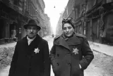 A man and a woman wearing overcoats, each with a prominent yellow star, stand in a rubble-strewn street.