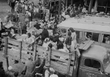 A black-and-white photograph of people being loaded onto trucks as others behind them wait in line.