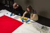 Two women wearing protective gloves and aprons measure the length of a large Canadian flag, which lies flat on a table.