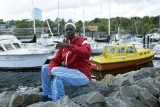A man wearing a brightly coloured track suit and a white skullcap sits on some rocks, with boats and a harbour in the background.