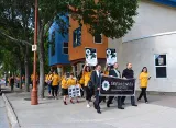A small crowd of people wearing yellow shirts march down a sidewalk, following three men in dark clothes who hold a banner reading “Srebrenica 11.07.1995.”