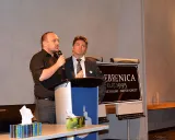 Two men stand at a lectern next to a sign reading “Srebrenica, July 11, 2015. Never Forget.”