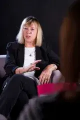 A woman sitting in a chair. Her left elbow is resting on the arm rest and her right hand is reaching in front of her as she is talking.