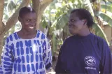Two women are standing in front of tropical trees, looking at each other and smiling. The woman on the right is wearing a sweater that bears the logo of the Concerned Parents Association.