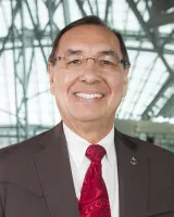 An indigenous man with short brown hair and glasses. He's wearing a dark grey hair and red tie.