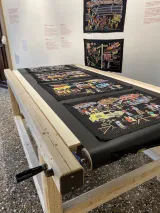 A series of colourful embroidery panels on black fabric displayed on a wooden hand-cranked conveyer belt table and on the wall of a museum.