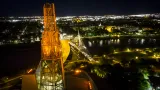 A glowing orange tower is pictured from above against a background night-time cityscape with view of a river and a bridge.