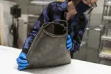 A woman holds a scuffed black rubber bag with a roughly shaped curved metal handle spanning the top.