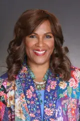 A woman with dark hair, dark eyes is smiling with a multi-coloured top on.