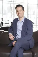 A caucasian man in a business suit has dark hair and blue eyes. He is sitting cross-legged.