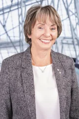 A white woman with light brown hair is wearing a grey patterned blazer and a white shirt. She is smiling facing the camera and standing in a relaxed manner.