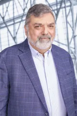 A man with grey hair wears a blue-purple blazer with a white shirt.