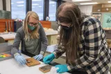 Two people wearing face masks and blue gloves stand at a white worktable and use small swabs to clean pieces of wood covered with photographs and text.