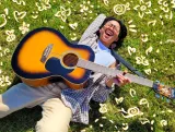 Kris Cahatol lies laughing on green grass and is surrounded by computer-drawn flowers and hearts. Kris is holding a guitar across their chest.