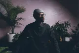 Alpha Toshineza, a Black man wearing a toque, round glasses and an intense expression, stands in a room full of tropical plants.