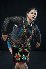 Melody McKiver, Anishinaabe, porte un chemisier noir transparent sur un débardeur arc-en-ciel, du rouge à lèvres noir et une expression intense. Iel se penche vers l’avant, ses cheveux noirs en deux tresses, avec un carré de tissu brodé porté en bandoulière sur une lanière perlée.