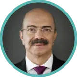 A man with a dark moustache and small, frameless glasses wears a dark suit and a purple tie.