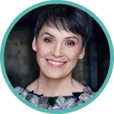 A woman with short dark hair and a blouse with a collar of flowers smiles.