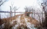 A rusted gate closes off a narrow track that vanishes around a bend. Large bodies of water are visible on both sides of the grassy road. 