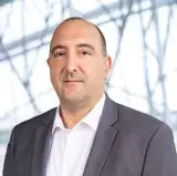 A white man wears a dark grey blazer with a white dress shirt. He is facing the camera and has a slight smile.