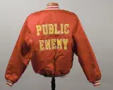 The back of a deep orangey-red bomber jacket decorated with “Public Enemy” in large yellow capital letters.