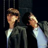 Two people wearing white shirts and black blazers pose against a black-painted cinderblock wall.