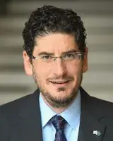 White man with short dark hair and glasses looks ahead with a smile. He's wearing a tie and jacket.