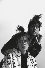 Two people with long hair in high ponytails. One stands behind the other and leans on their shoulders, wearing a black and white patterned shirt under a black leather jacket. In front, the other wears a solid black shirt under a black-and-white patterned jacket. 