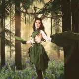 A drag artist is dressed in a cosplay costume with long, brown hair; large antlers; and green leaf scarf, skirt and gloves.