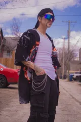 A drag artist is wearing a black cap, shirt and pants with chains, as well as blue shades and a white tank top.