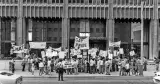 A group of people protesting on the sidewalk in front of an office building. They are holding protest signs and banners and are arranged three and four deep. A police officer watches them.