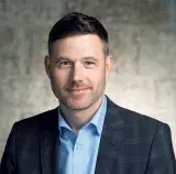 A white man in a checked suit jacket and blue shirt is posing for a professional headshot against a stone background. He has short dark hair and a trimmed beard.