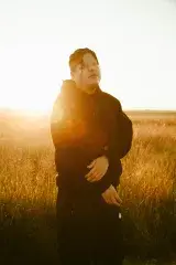 An Indigenous man with short, black hair wearing a black hoodie and pants is in a golden wheat field with a sunset in the background.