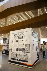 Exhibit in a modern space showcasing historical posters and text about 2SLGBTQ+ activism, with people observing the display and a person on a balcony above.