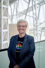 A woman with light hair and dark glasses smiles at the viewer. She is wearing a dark suit jacket and t-shirt that features a map of Canada in the colours of the inclusive Pride flag.