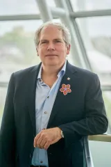 A man stands facing the viewer with a serious expression. He is wearing a dark suit jacket and a light blue buttoned-up shirt. He sports a multicoloured Métis-style flower on his lapel.