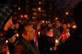 A large group of people stand together outdoors at night. Many are holding candles.