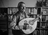 A man in a checkered shirt and vest plays a large stringed instrument with bookcases filled with books in the background.