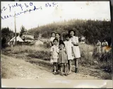 Black-and-white image of women and children standing on a gravel road in front of a field. 