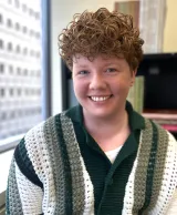 A white person with short curly dark blond hair smiles into the camera. They are wearing a loose, lightweight sweater with vertical white, grey and black stripes.
