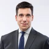 A white man with dark hair smiles slightly into the camera. He is wearing a dark suit with an off-white shirt and a dark blue tie with small white polka dots.