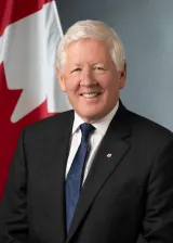 A white man with grey hair stands smiling in front of a Canadian flag. He is wearing a black suit with a blue tie and white shirt.